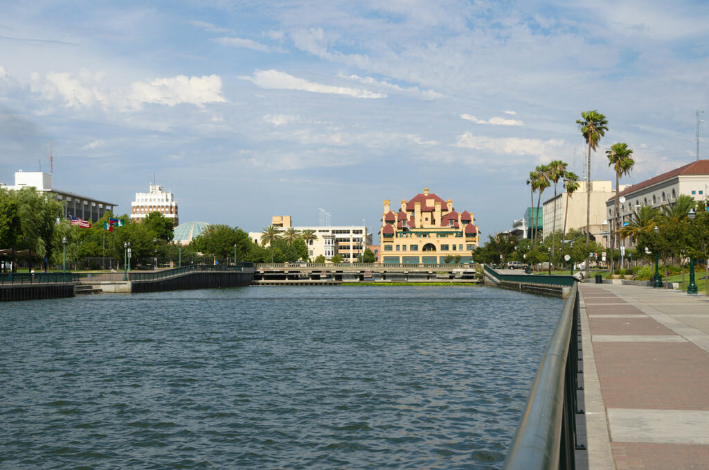 A wide river runs alongside a paved walkway bordered by trees and buildings under a partly cloudy sky, perfect for any pet lover enjoying a stroll with their furry friend.